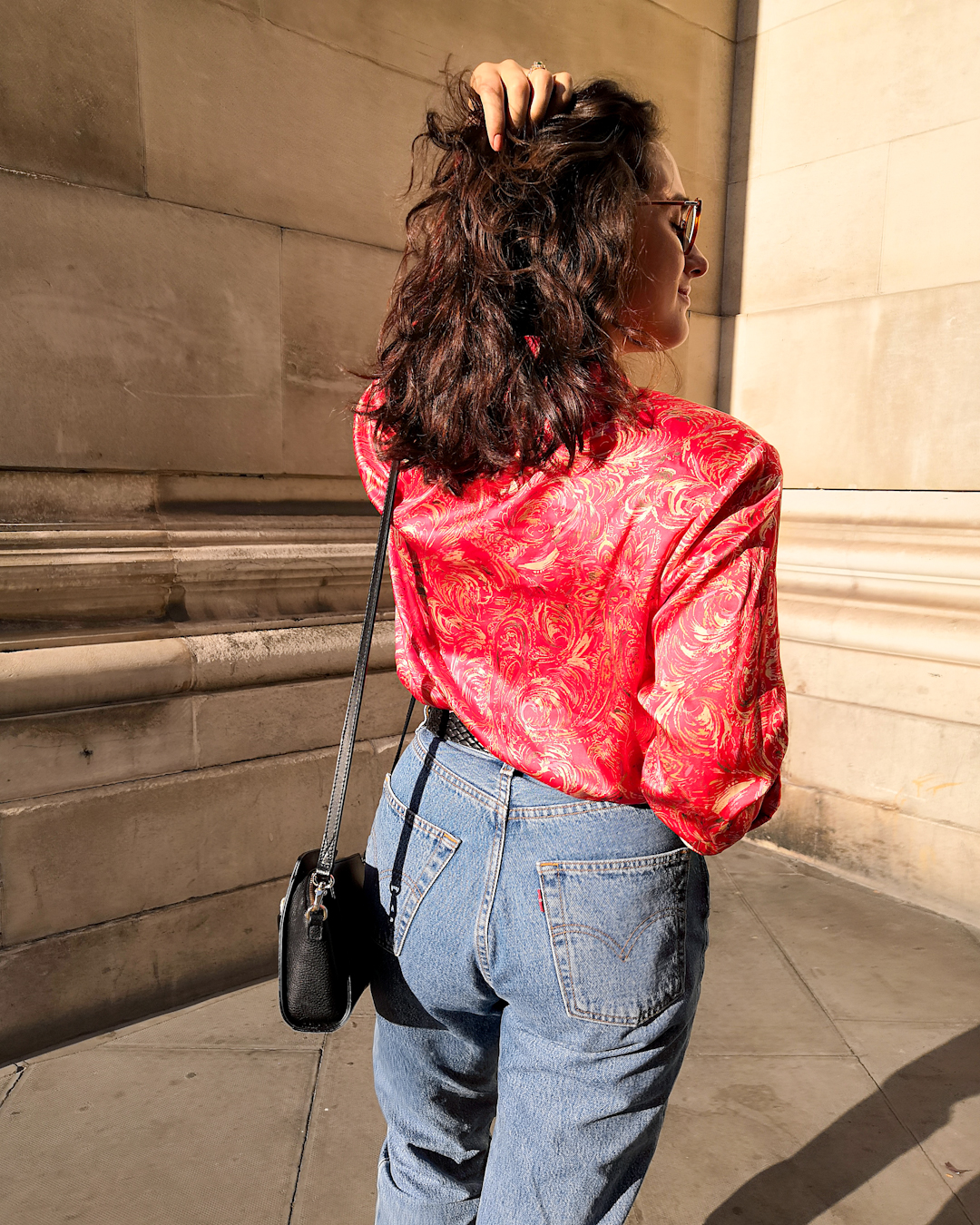 Katie wears a red and gold patterned blouse with high waisted denim cuffed above the her Dr Martens boots. Her hair is down and wavy. She's standing in front of a stone wall with minimal details.