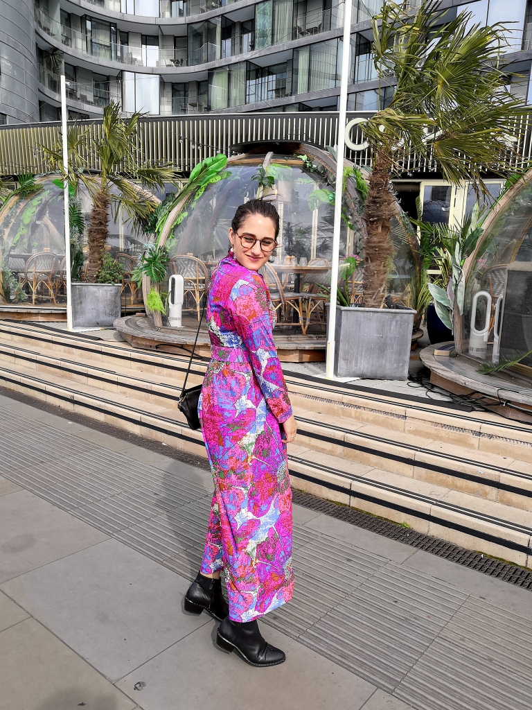 Katie wears a pink, blue and white multi colour vintage maxi dress with long sleeves, high neck, matching belt and black shoes and a small black shoulder bag. Her hair is pulled back in a pony tail and she's standing in front of the surfer shacks at Copa Club on Tower Bridge.